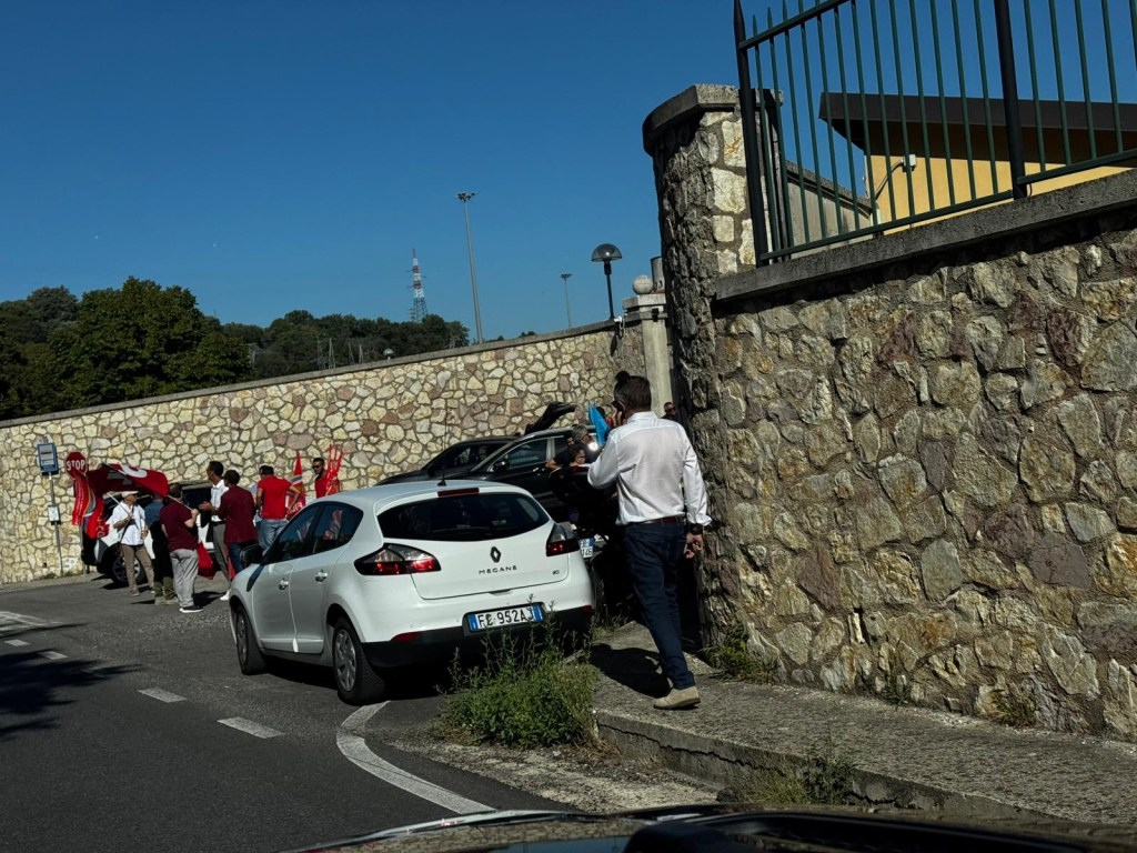 Un gruppo di persone con bandiere rosse protesta davanti a un edificio, con alcune auto parcheggiate in strada.