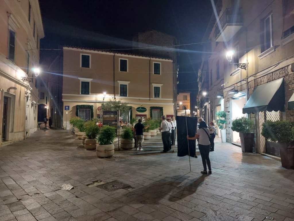 A night scene of a quaint street with people gathered, illuminated by street lamps, showcasing a blend of historic architecture and outdoor plants in pots.