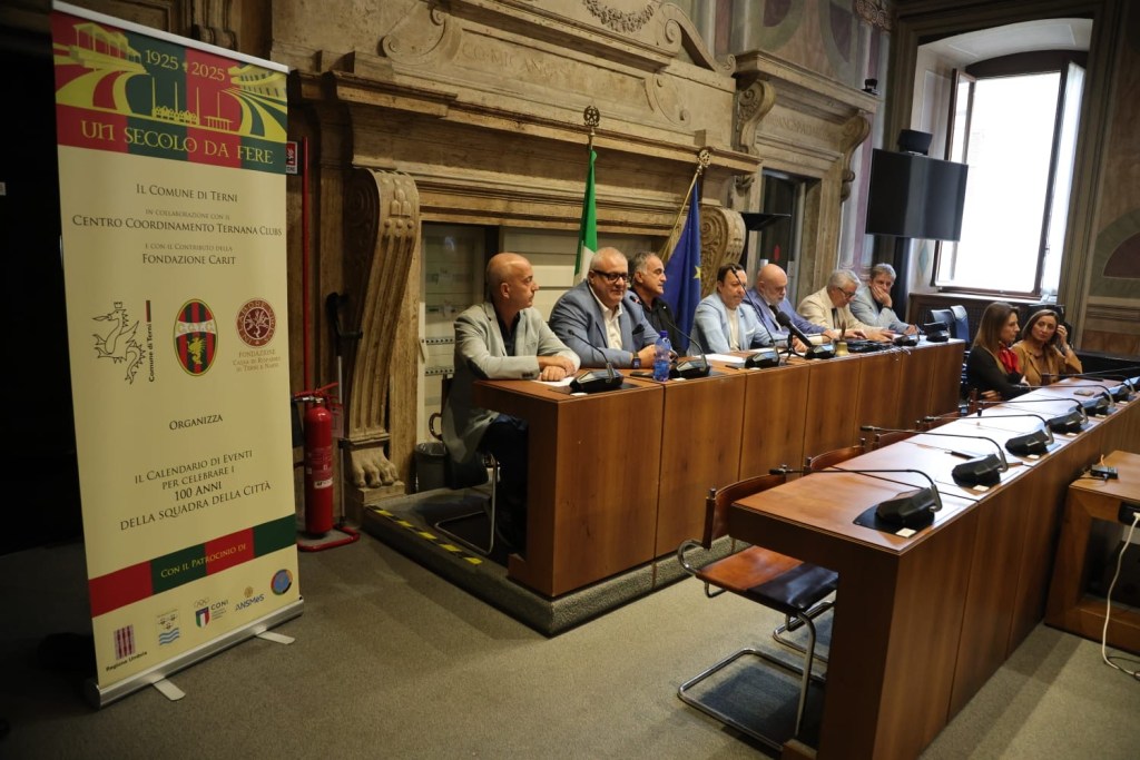 A panel of speakers seated at a long table during an event, with a banner displaying information about a centenary celebration in the background. The setting appears to be an official conference room with decorative architecture.