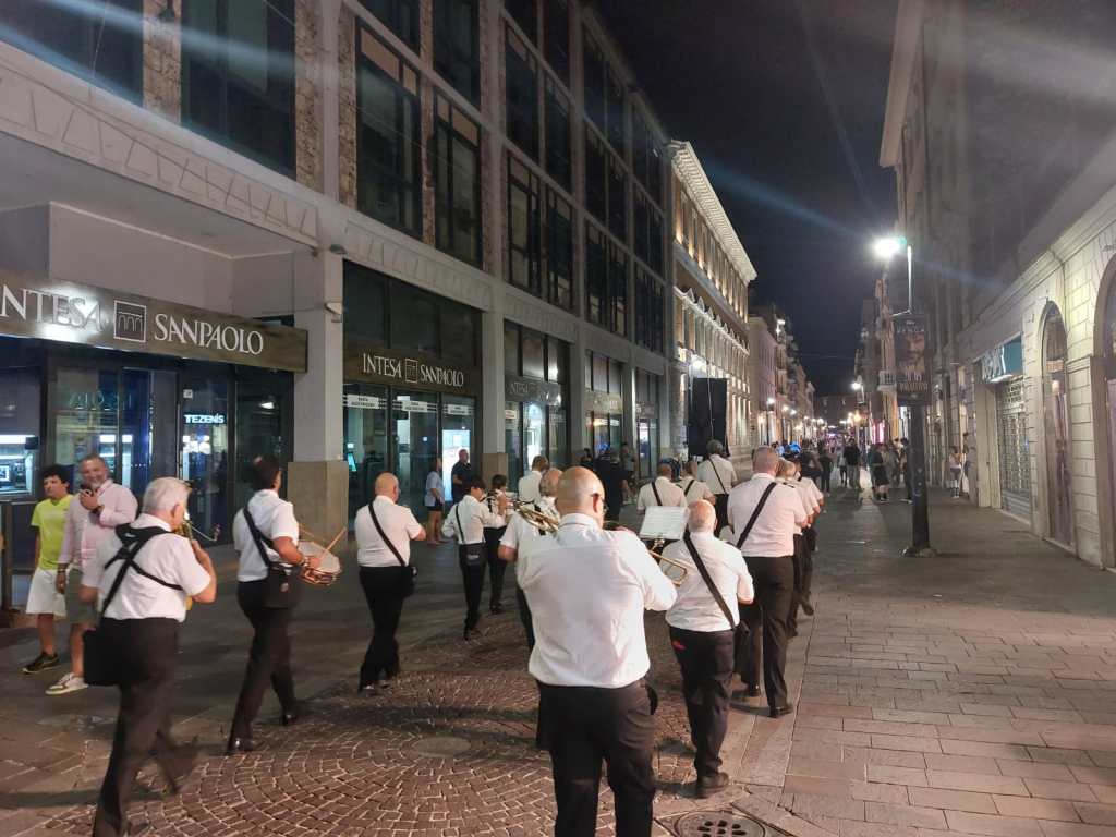 A street scene at night featuring a group of musicians wearing white shirts and black pants, playing instruments while walking along a cobblestone street lined with shops and people.