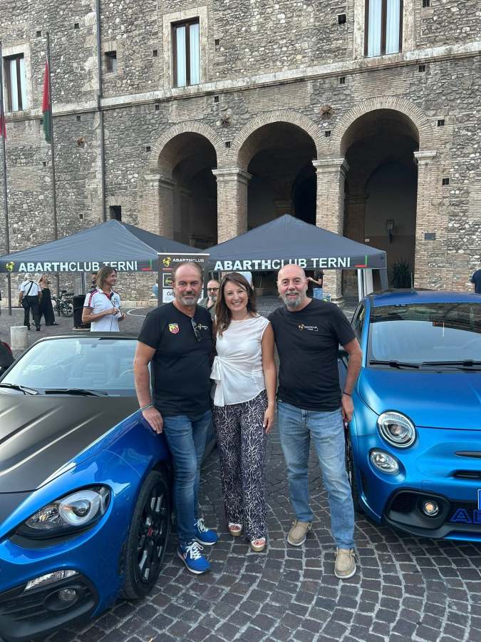 Tre persone, due uomini e una donna, sorridono davanti a due auto sportive blu, in una piazza di Terni. Sullo sfondo, un edificio storico e tende con il logo di Abarth Club Terni.