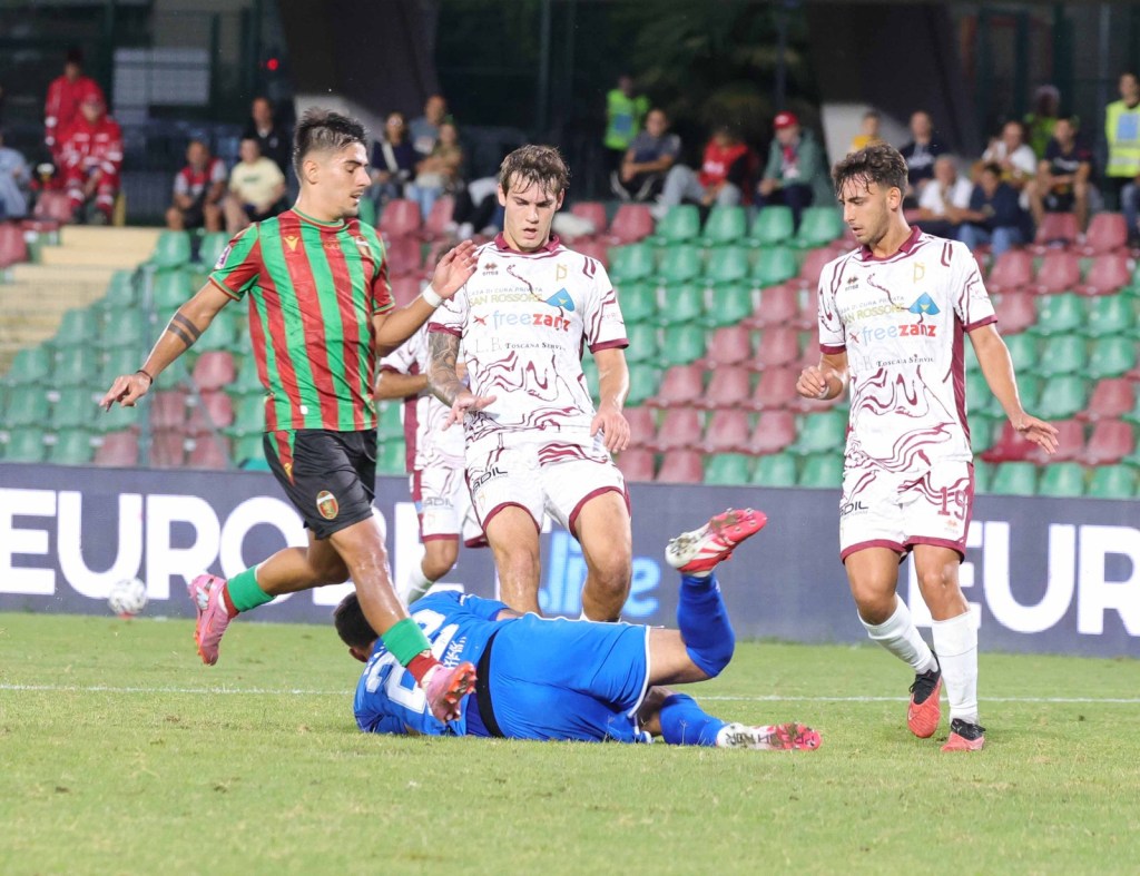 A soccer match in progress, featuring players in red and green uniforms competing with those in white. One player is in a kneeling position, while others are engaged in a play.