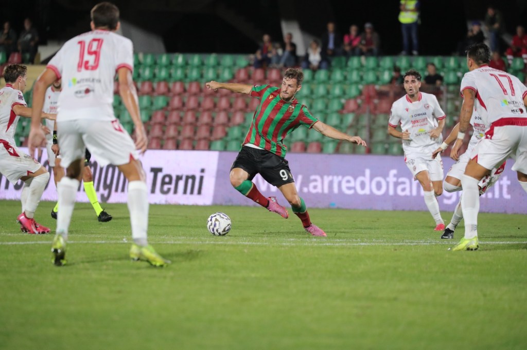 A soccer player in a red and green striped jersey dribbles the ball while being surrounded by opposing players in white jerseys during a match.