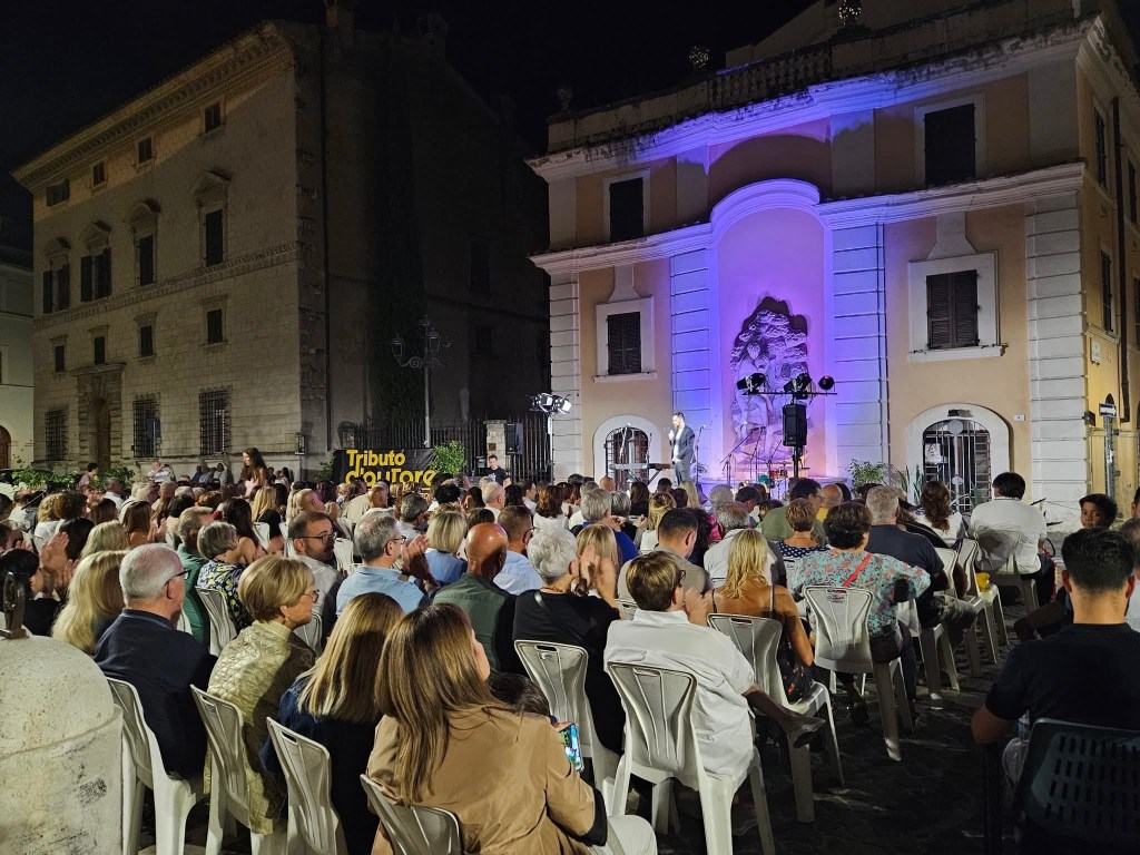 A lively outdoor event with a large audience seated in plastic chairs, facing a stage illuminated with purple lights. The backdrop features a decorative wall, and the atmosphere is festive at night.