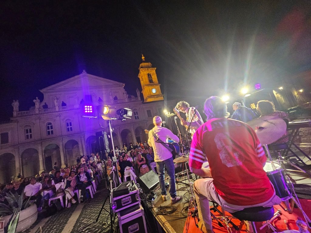 A live music performance in a vibrant plaza at night, featuring musicians on stage and a large audience enjoying the show. The backdrop includes a historic building with illuminated clock tower.