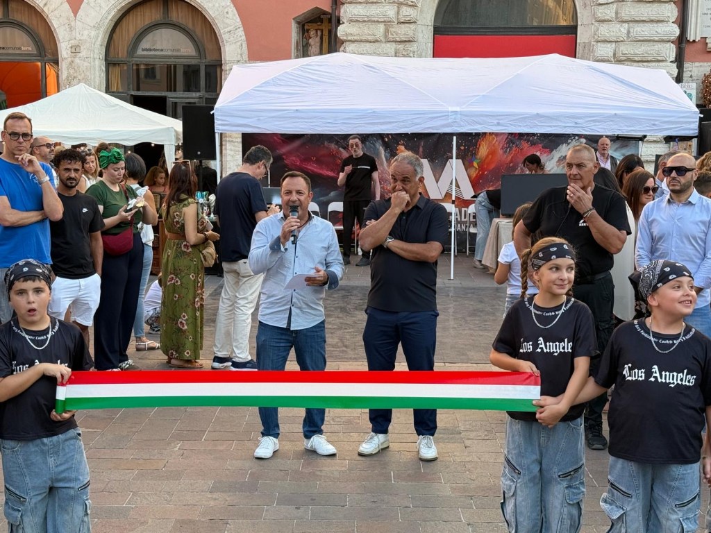 A speaker addresses a crowd at an outdoor event, while children holding a ribbon stand in front. Various attendees can be seen in the background.