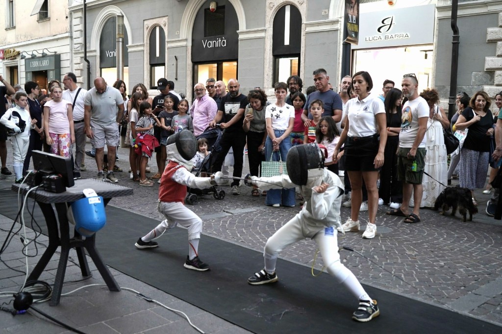 Two children fencing in a public square, with a crowd of onlookers watching the event. The setting includes a backdrop of shops and a mix of adults and children in the audience.