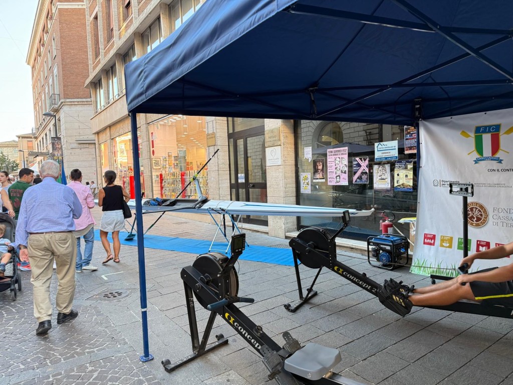 A street scene featuring a promotional booth with rowing machines set up on a sidewalk. People are walking by, and a participant is using one of the rowing machines. The booth is adorned with banners related to rowing, and a variety of businesses can be seen in the background.