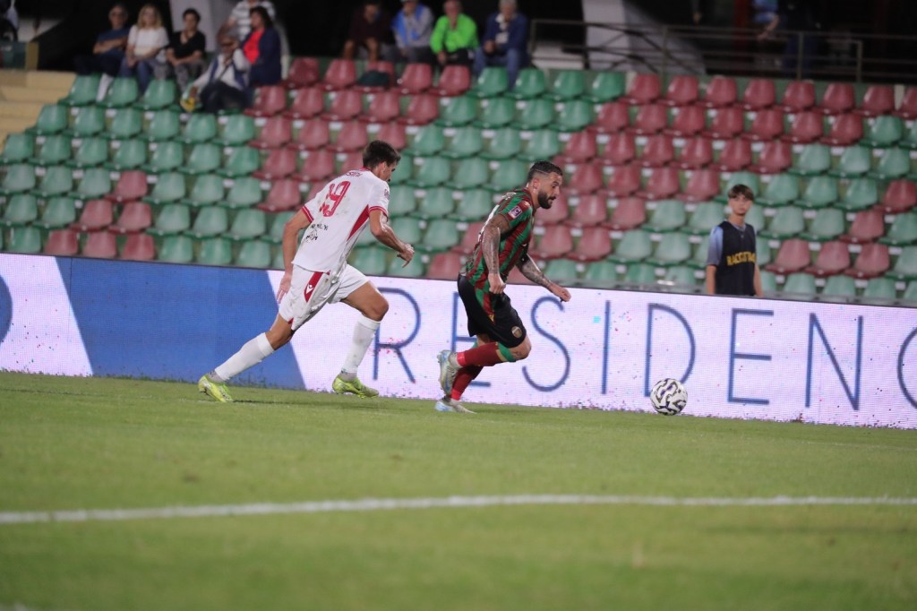 Two soccer players competing for the ball on a grassy field with a partially filled stadium in the background.