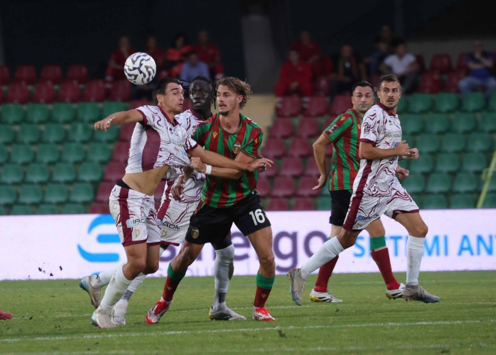 Giocatori di calcio in azione durante una partita, con un atleta che tenta di colpire il pallone mentre un avversario lo ostacola.