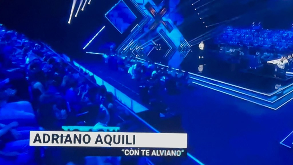 Concert scene featuring a stage with blue lighting and an audience. A performer named Adriano Aquili is on stage, with the title 'Con Te Alviano' displayed.