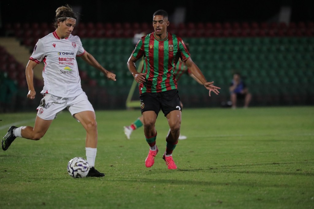 A soccer match in progress, featuring two players in action on a grass field. One player in white is dribbling the ball while another player in a green and red striped jersey is defending.