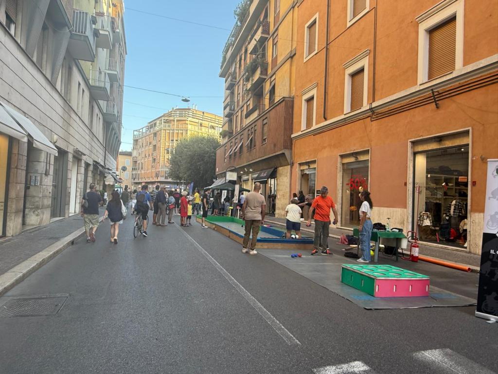 A vibrant street scene in a city, featuring people walking and engaging in activities. Colorful mats and equipment are set up on the road, indicating a community event. Buildings with warm-colored facades line the street, complemented by a clear blue sky.