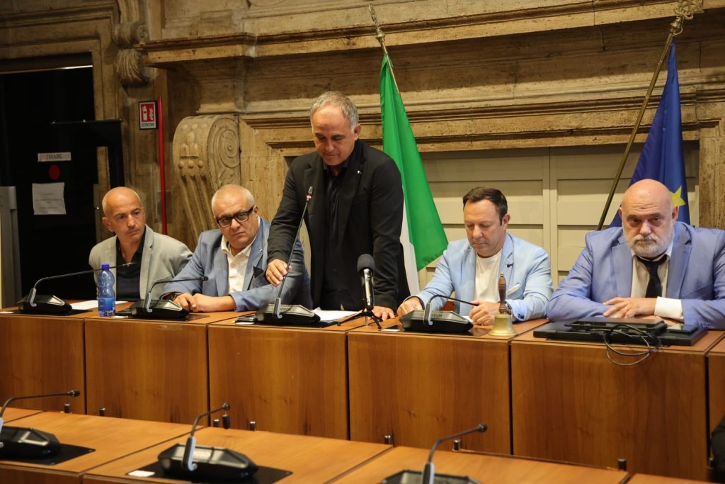 A group of five men sitting at a conference table, with microphones and documents in front of them, in a formal setting. The backdrop features a green Italian flag and a blue European Union flag.