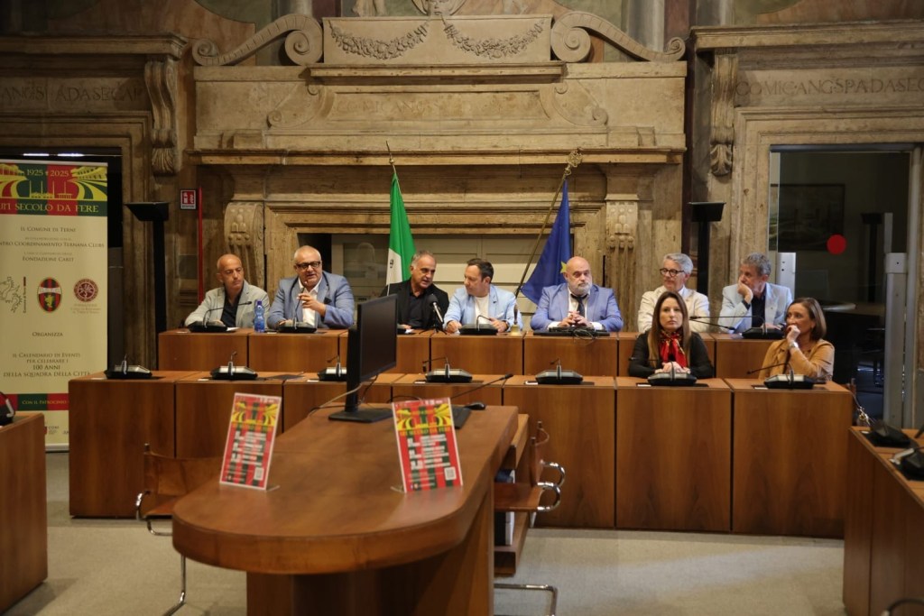A meeting in a formal setting with several speakers seated at a long wooden table, an Italian flag and a blue flag in the background, discussing an event related to the city's history.