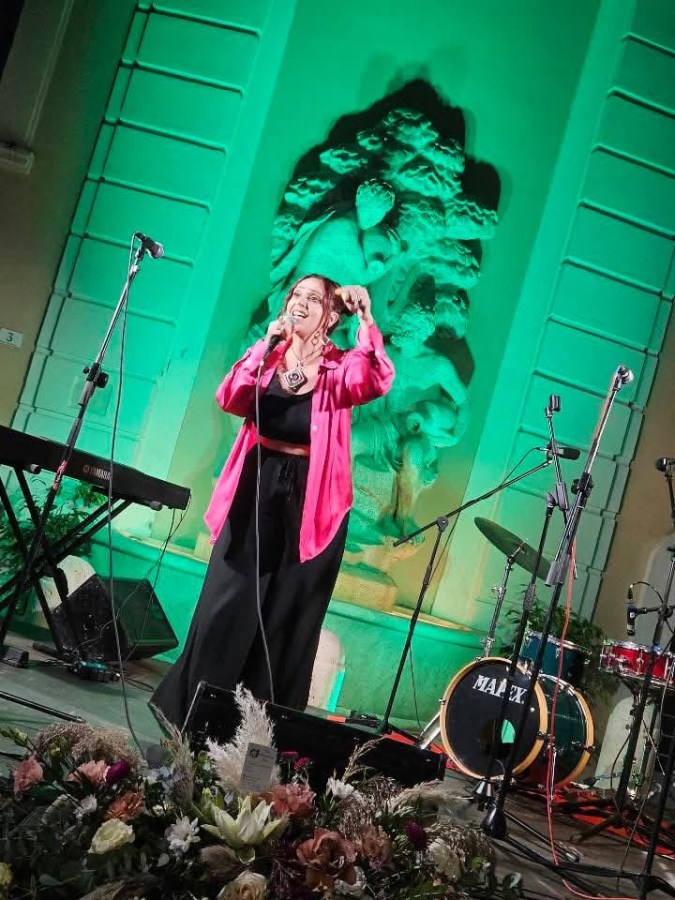 A female performer sings passionately on stage, wearing a bright pink blouse and black pants. In the background, a green-lit decorative wall and musical instruments are visible, including a keyboard and drums. The stage is adorned with a floral arrangement in the foreground.