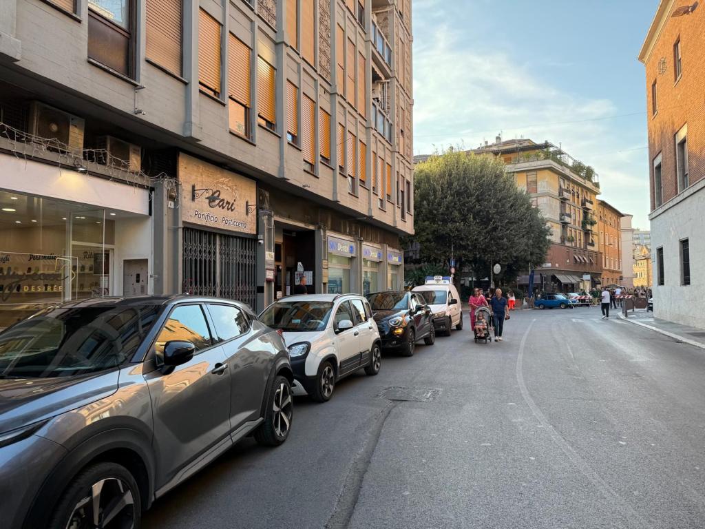 Street scene with parked cars along a narrow road, featuring a bakery named 'Pani' and pedestrians walking, set against a backdrop of buildings and clear blue sky.