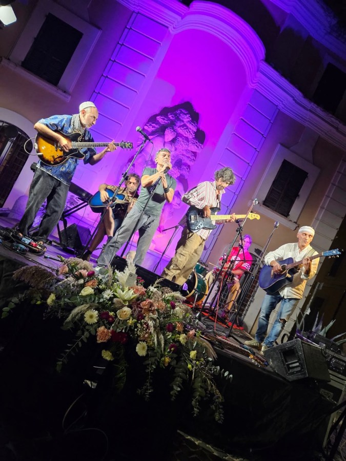 A live music performance featuring five musicians on stage with various instruments, illuminated by colorful lights, in front of a floral arrangement.