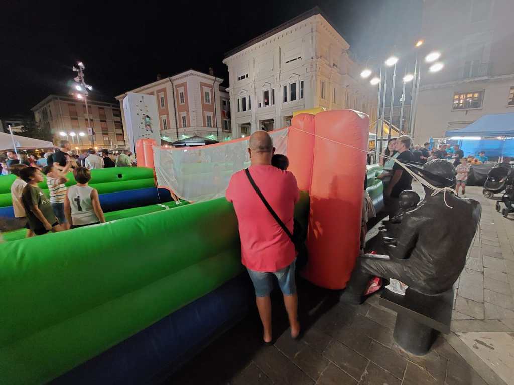A lively night market scene with people gathered around inflatable structures. A person in a pink shirt is observing from behind a green inflatable barrier, while children play nearby. Festive lights illuminate the area.