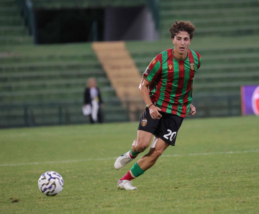Un giocatore di calcio in maglia verde e rossa corre sul campo, inseguito da una palla da calcio.