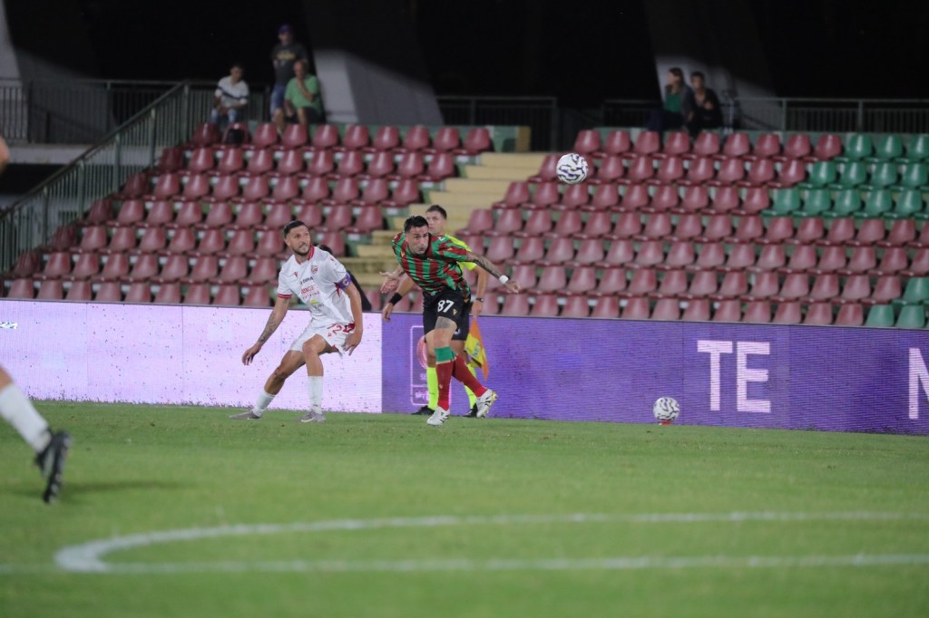 A soccer match in progress, with players from opposing teams on the field. A player in a colorful jersey is about to kick the ball while a teammate in white watches. The stadium seating is partially filled.