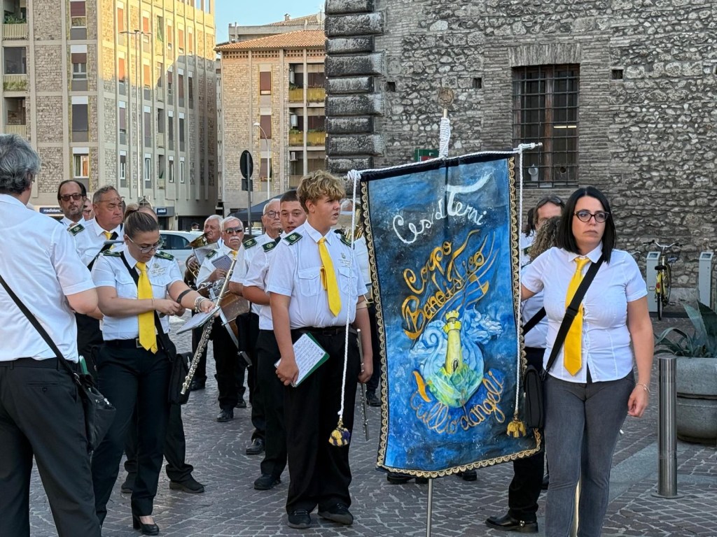 A group of musicians in white shirts and yellow ties prepare for a performance, holding instruments and a colorful banner in a public square.