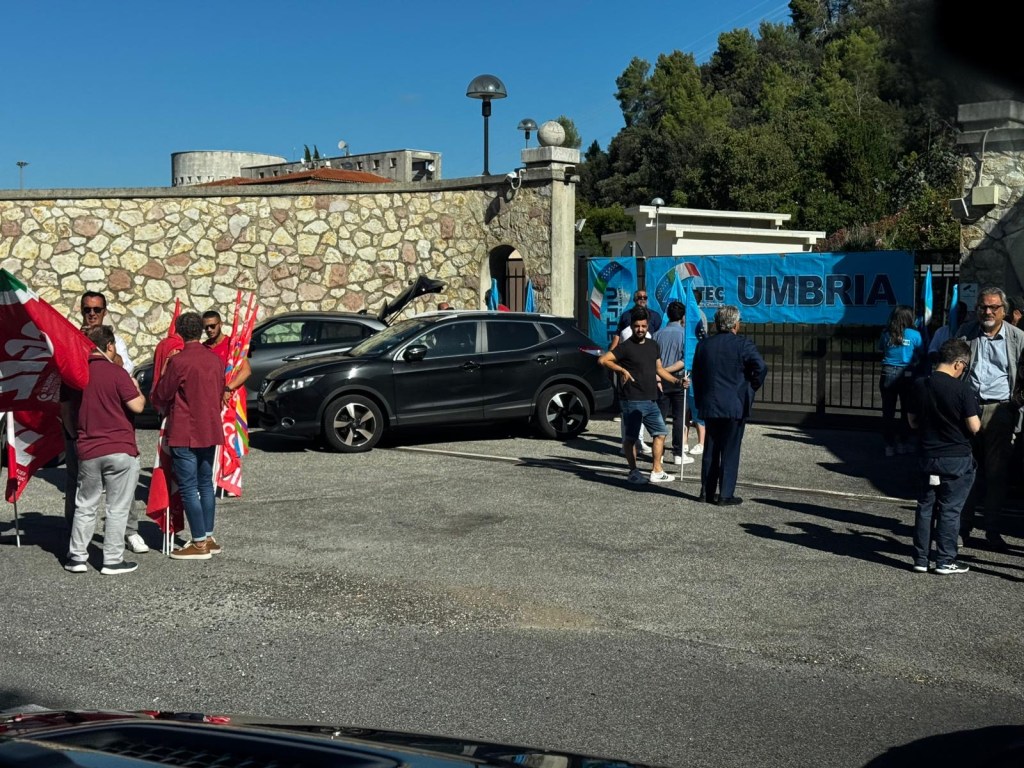 Manifestazione di lavoratori e cittadini davanti a un cancello, con bandiere e striscioni per protestare contro la chiusura del posto di teleconduzione di Terni.