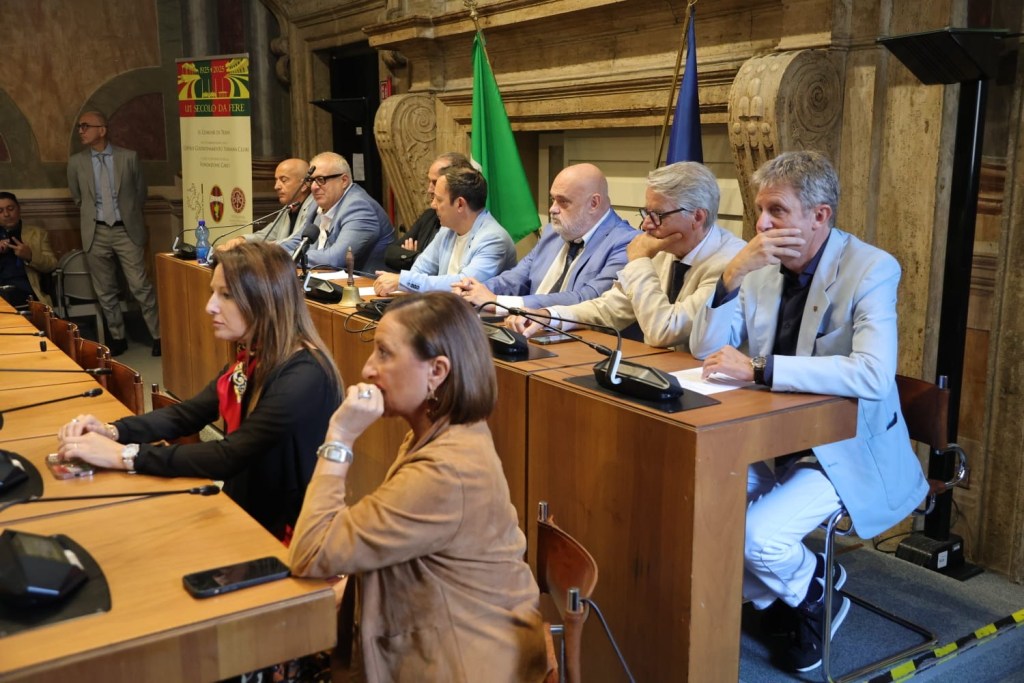 A group of people seated at a long table during a meeting, with microphones and water bottles in front of them. In the background are flags and a banner, with a partially visible wall art.