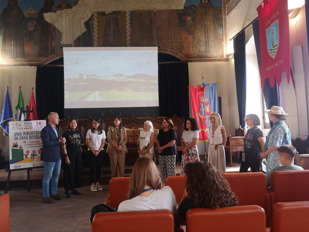 Evento di premiazione con relatori e partecipanti in una sala decorata, con uno schermo e bandiere sullo sfondo.