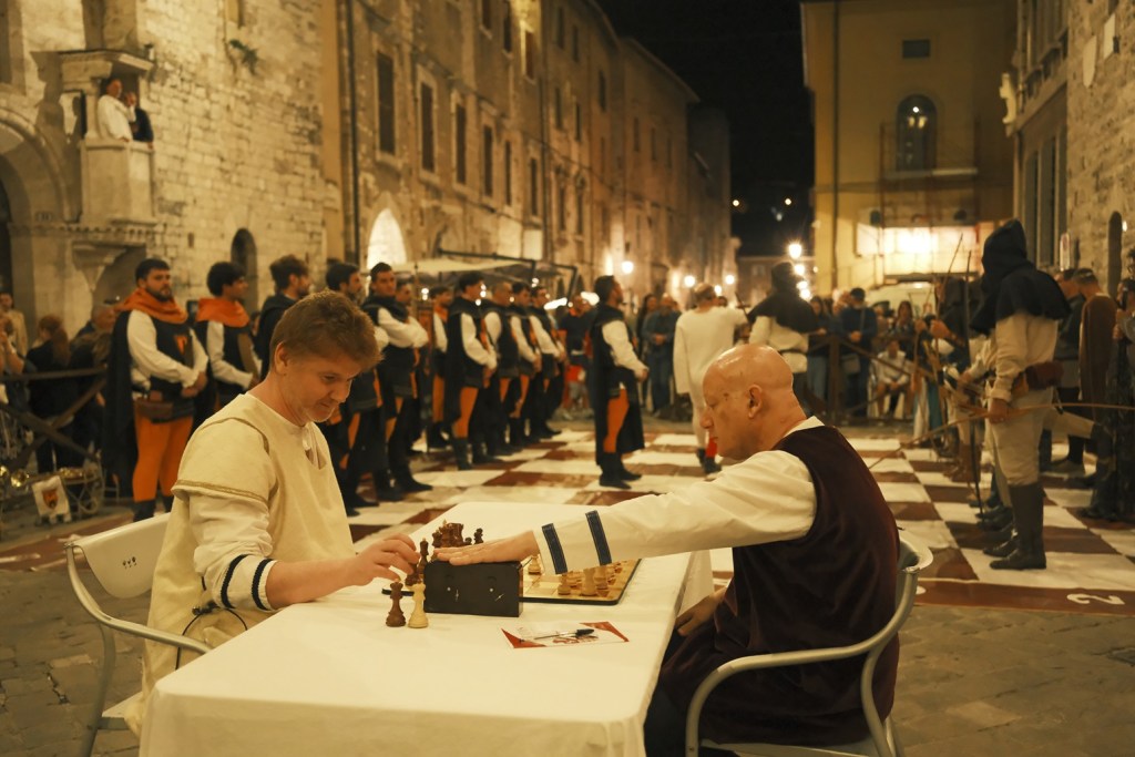 Due uomini giocano a scacchi su una scacchiera gigante durante il Festival delle Arti del Medioevo a Narni, con un pubblico in costume medievale che osserva sullo sfondo.