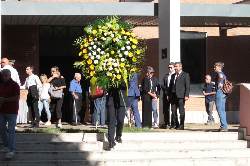 A person carries a large floral arrangement while a group of people stands nearby, some watching attentively. The scene is set outside a building with a modern architectural style.