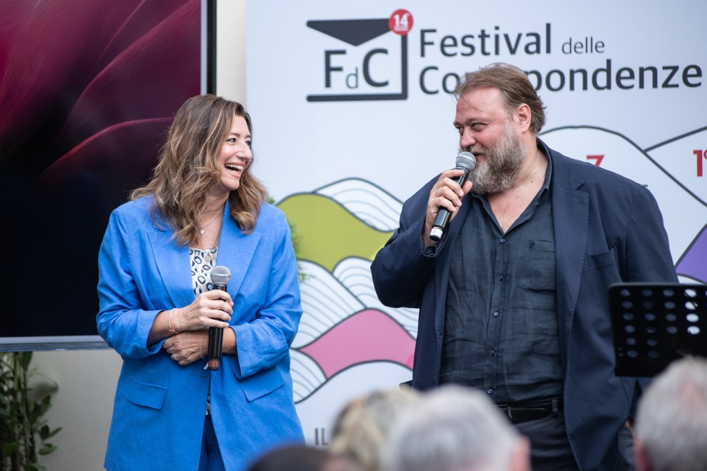 Due relatori sorridenti, uno in giacca blu e l'altro in giacca scura, conversano sul palco durante il Festival delle Corrispondenze, con uno sfondo colorato e il logo dell'evento.