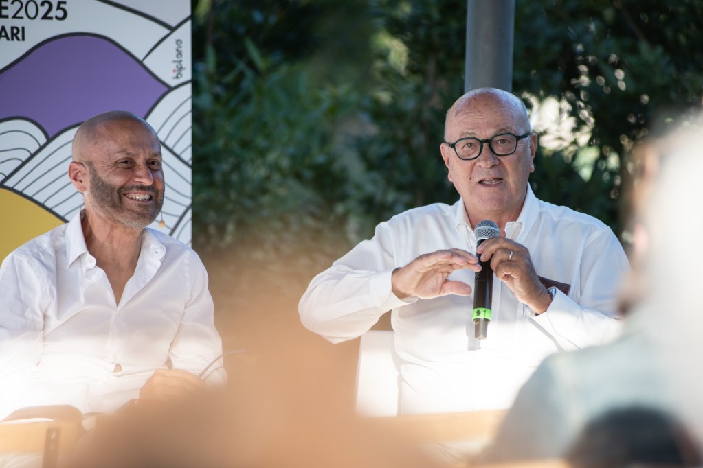 Due relatori seduti a un tavolo con un microfono, entrambi sorridenti durante un evento del Festival delle Corrispondenze a Magione, Umbria.