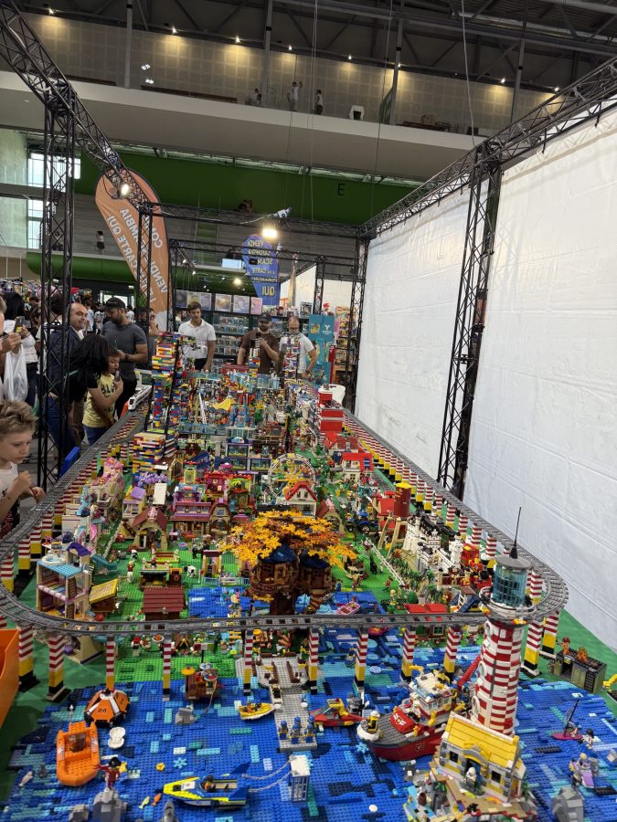 Un grande diorama realizzato con mattoncini LEGO, che rappresenta una vivace città con case, un albero, una torre faro e diversi veicoli, circondato da visitatori interessati all'esposizione.