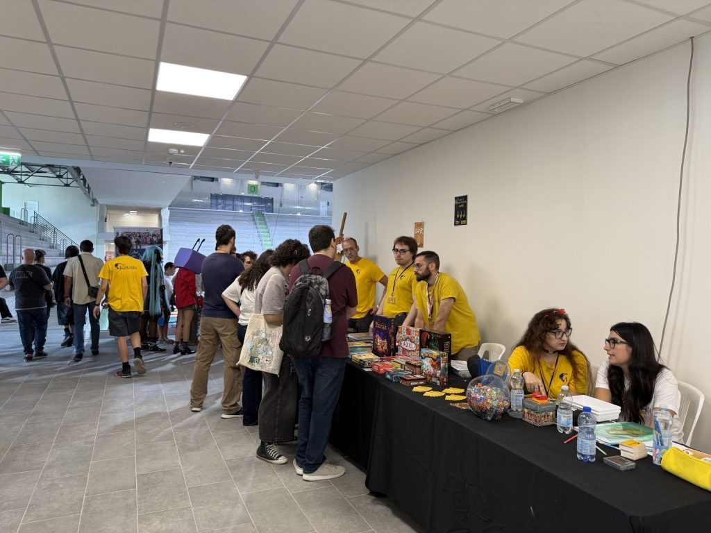 Un gruppo di persone interagisce al bancone di un evento, circondato da tavoli con giochi da tavolo e carte. Alcuni partecipanti indossano magliette gialle.
