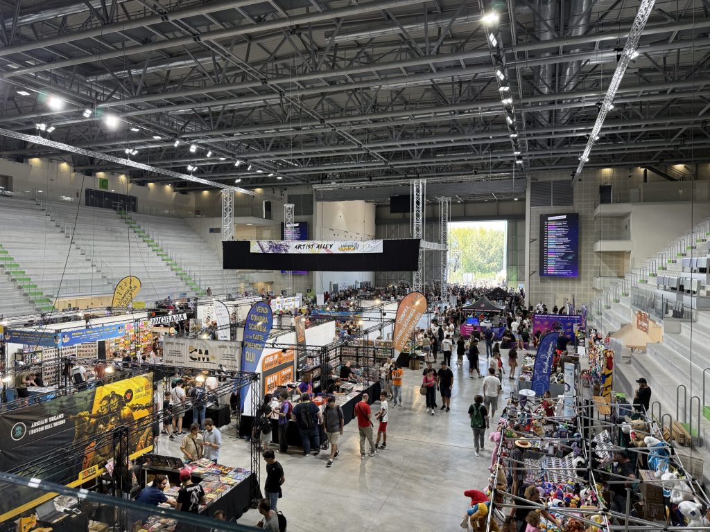 Panoramica di un'area interna del Palaterni durante Terni Comics, affollata di visitatori che esplorano stand di fumetti, giochi e merchandise, con elementi decorativi e banner colorati.