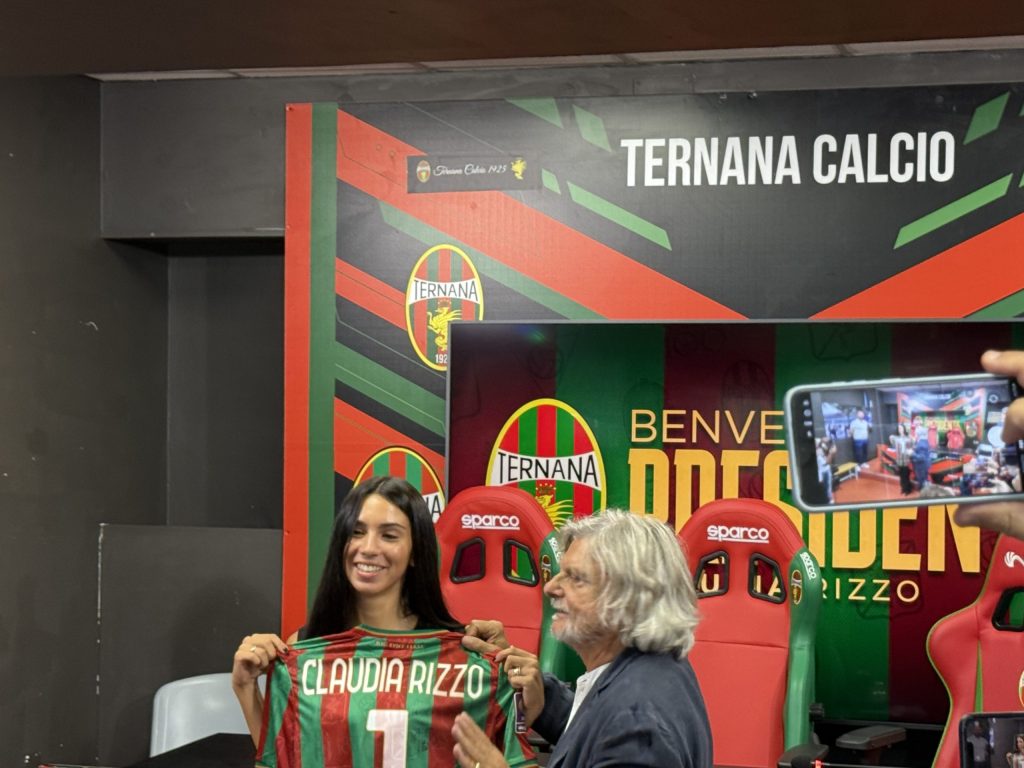 Claudia Rizzo holds a Ternana Calcio jersey with her name and number during her first press conference as president, surrounded by supporters and staff.
