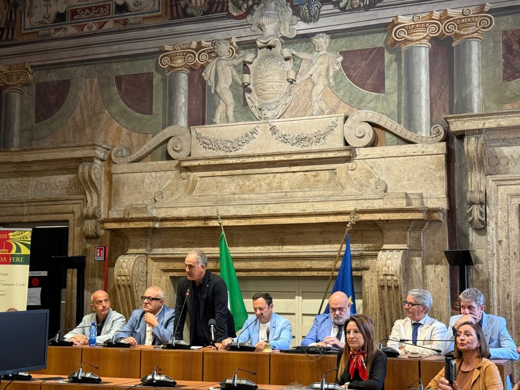 A panel discussion taking place in a formal setting, featuring multiple speakers at a long table, with decorative architecture in the background, including flags.