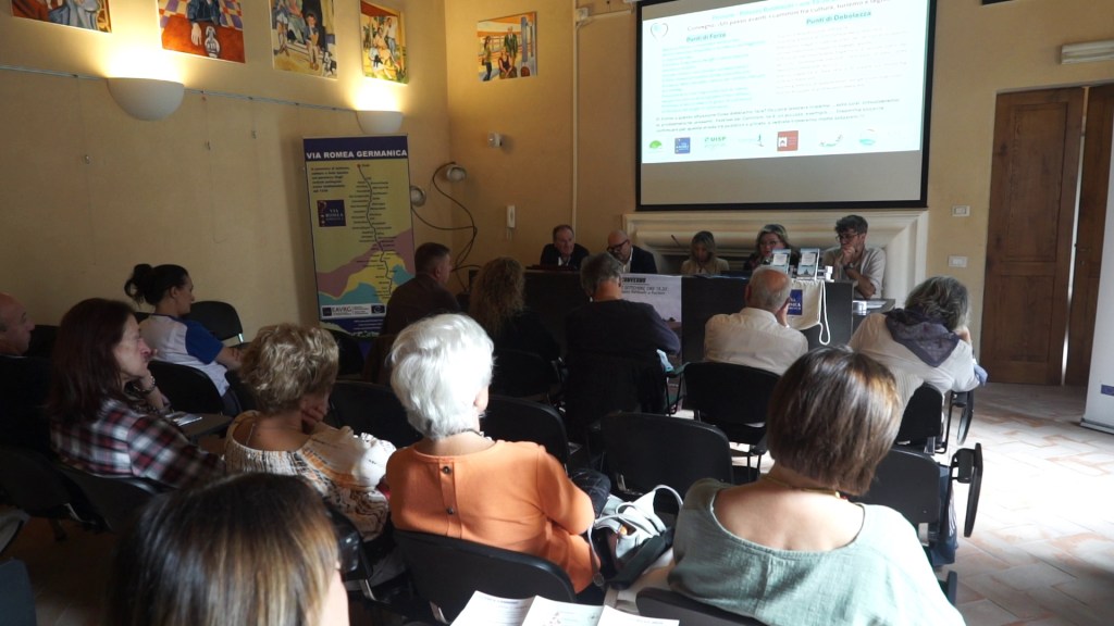 A conference on the promotion of walking paths held in Paciano, featuring a panel of speakers and an audience engaged in discussion.
