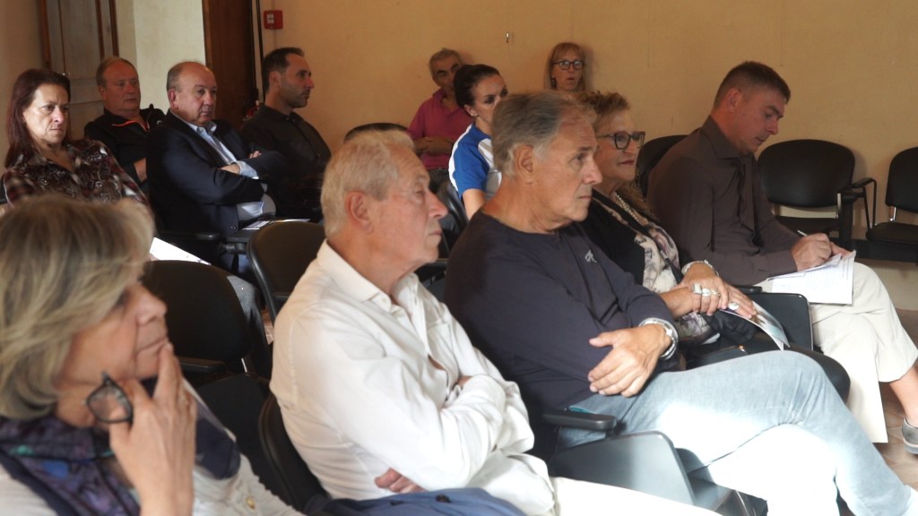 Un gruppo di persone ascolta attentamente durante un convegno, seduti su sedie in una sala. Alcuni partecipanti prendono appunti.