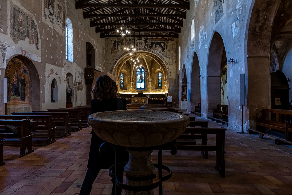 Interno di una chiesa storica con affreschi murali e un fonte battesimale in primo piano.
