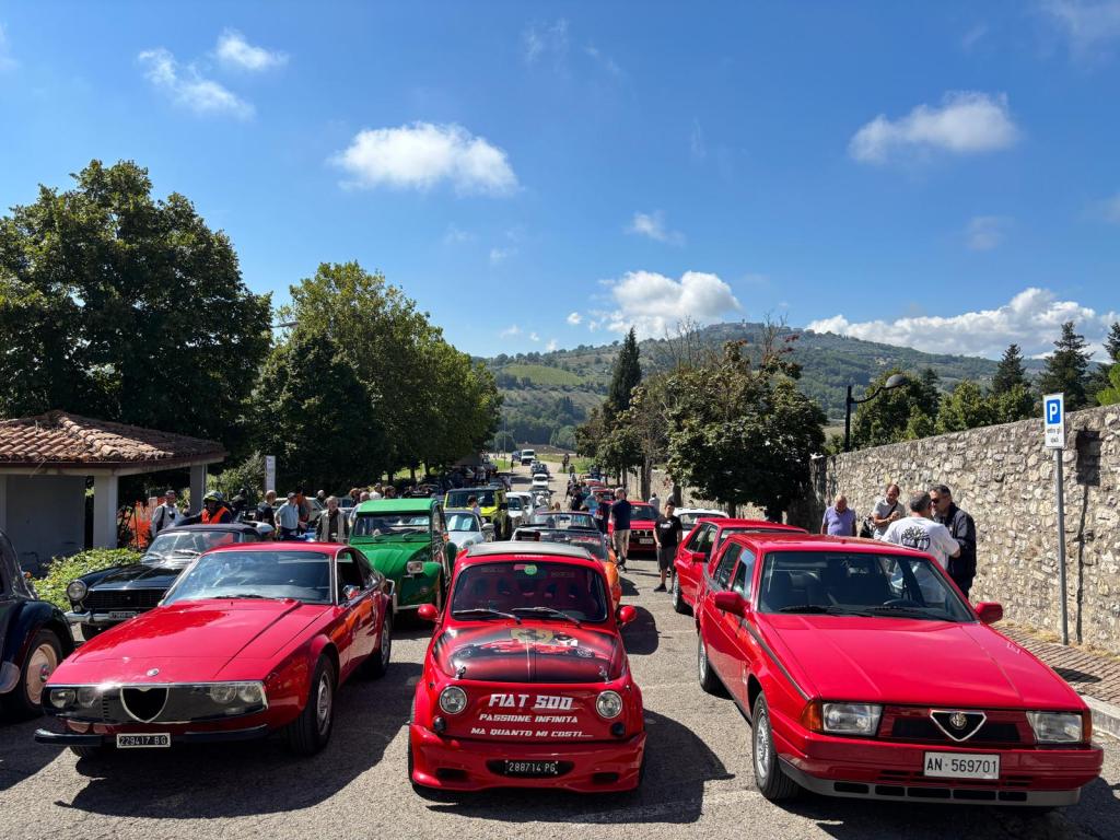 A gathering of vintage cars lined up on a street during a classic car and motorcycle rally in Fratta Todina, surrounded by trees and a blue sky.