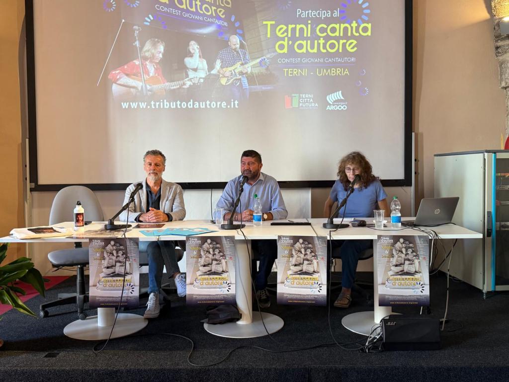 Three speakers at a table discussing the Terni canta d'autore contest, with promotional posters displayed in front of them.