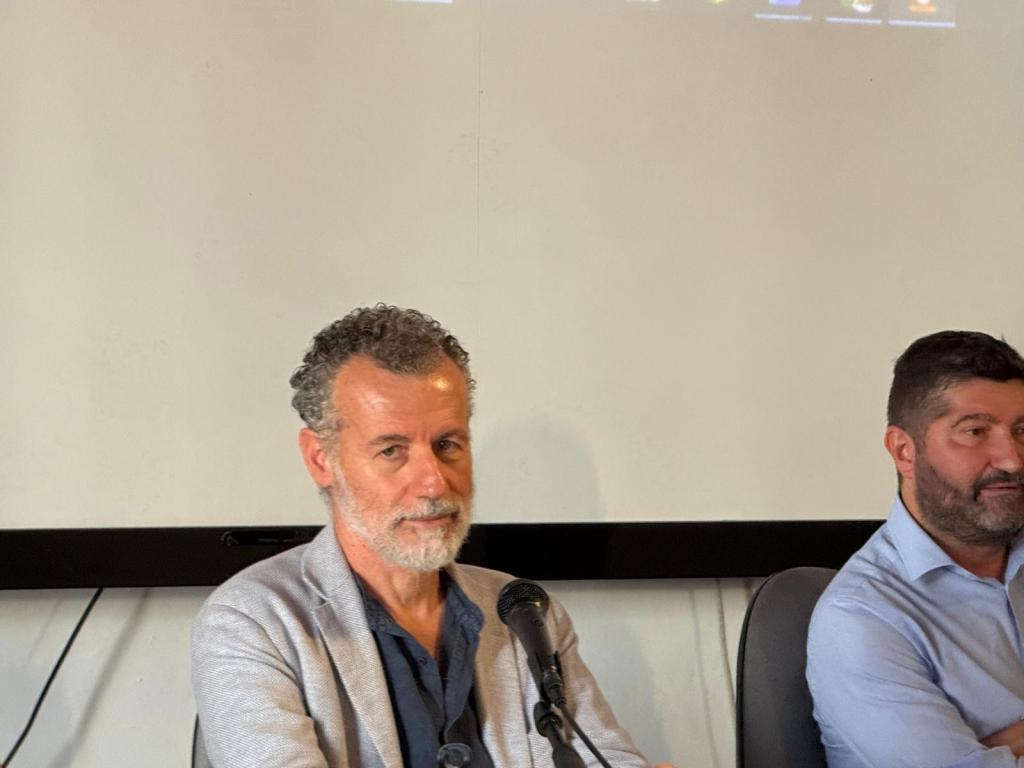 Two men seated at a table during a panel discussion, with one man in a light gray blazer and the other in a light blue shirt, both looking at the camera.