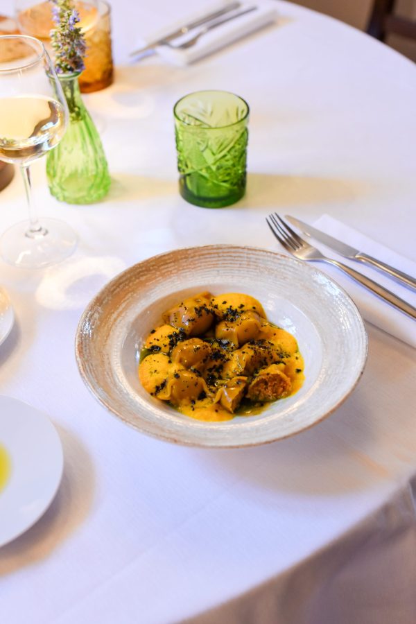 Un piatto di pasta fresca servito in un elegante piatto con salsa gialla e guarnizioni scure, accompagnato da bicchieri e posate su un tavolo apparecchiato.