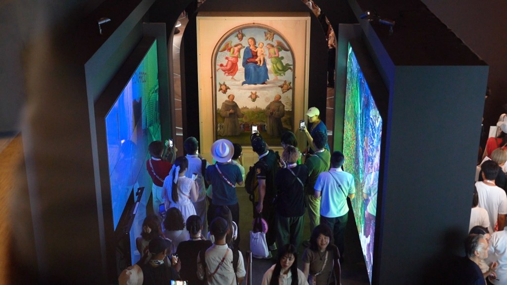 Un gruppo di visitatori osserva l'opera 'Gonfalone della giustizia' di Pietro Vannucci, esposta al Padiglione Italia durante l'Expo 2025 Osaka, mentre altri schermi colorati adornano l'ambiente circostante.
