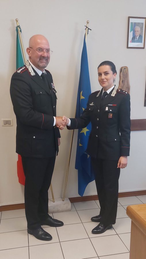 Two military officers, a man and a woman, are smiling and shaking hands in an office setting. The man is bald and wearing glasses with a uniform featuring insignia, while the woman has dark hair and is also in uniform. The background displays flags of Italy and the European Union.