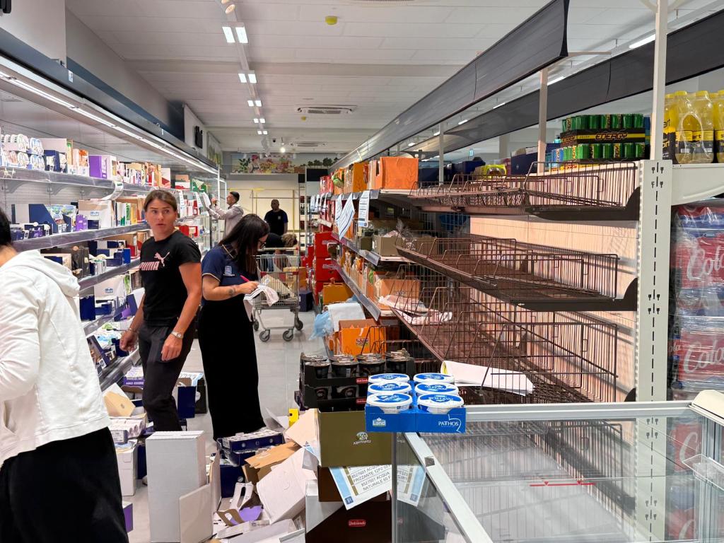 A busy supermarket aisle with shoppers and cluttered shelves, showcasing a partially restocked display and various products scattered on the floor.