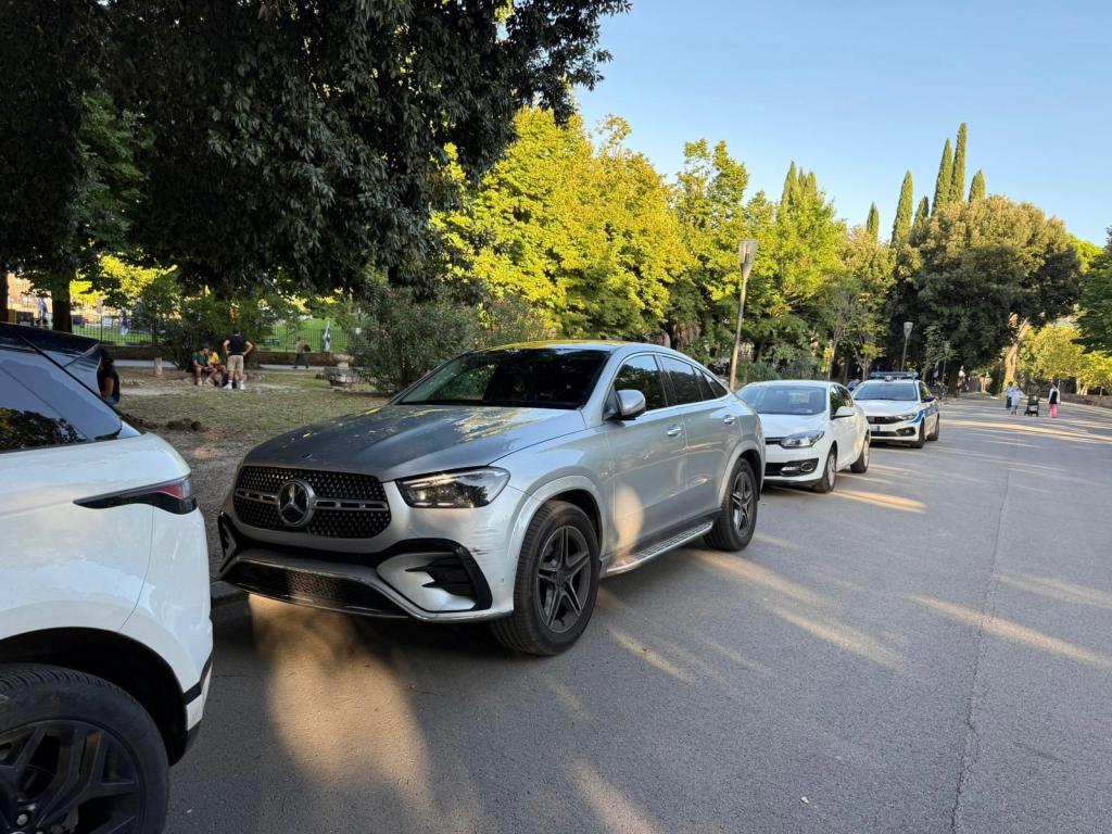 Una fila di automobilie in un parco monumentale, con un SUV Mercedes in evidenza. Gli alberi verdi circondano la strada.