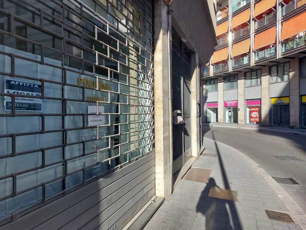 Vetrina chiusa di un negozio con serranda abbassata, affiancata da un cartello 'Affittasi' e altri negozi visibili sullo sfondo.